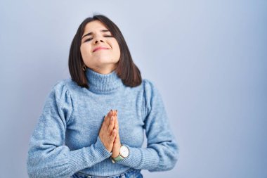 Mavi arka planda dikilen Latin kökenli genç bir kadın elleri bağlı yalvarıyor ve dua ediyor. Yüzünde umut ifadesi var. Çok duygusal ve endişeli. Yalvarıyor. 