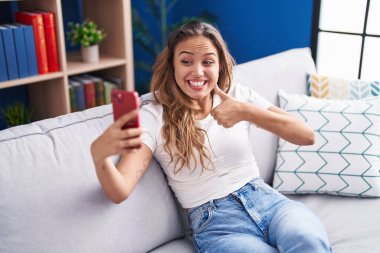 Young hispanic woman doing video call with smartphone smiling happy and positive, thumb up doing excellent and approval sign 