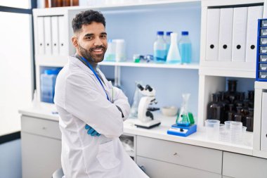 Laboratuvarda kollarını kavuşturup oturan bilim adamı üniforması giyen İspanyol genç adam.