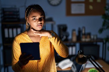 African american woman with braids working at the office at night with tablet cutting throat with hand as knife, threaten aggression with furious violence 