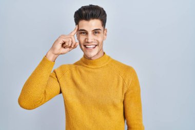 Young hispanic man standing over blue background smiling pointing to head with one finger, great idea or thought, good memory 