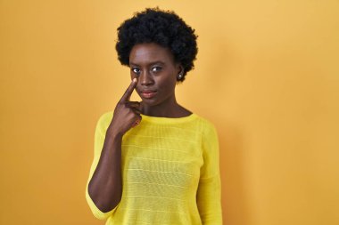 African young woman standing over yellow studio pointing to the eye watching you gesture, suspicious expression 
