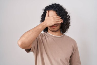 Hispanic man with curly hair standing over white background covering eyes with hand, looking serious and sad. sightless, hiding and rejection concept 