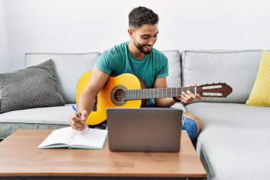 Genç Arap adam evde dizüstü bilgisayar ve klasik gitar kullanarak şarkı besteliyor.
