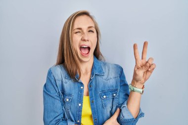 Young blonde woman standing over blue background smiling with happy face winking at the camera doing victory sign with fingers. number two. 