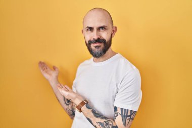 Young hispanic man with tattoos standing over yellow background inviting to enter smiling natural with open hand 