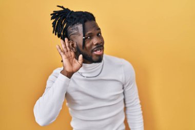 Sarı arka planda balıkçı yaka kazak giyen Afrikalı bir adam kulağına fısıldayarak dedikoduları ve dedikoduları dinliyor. Sağırlık kavramı. 