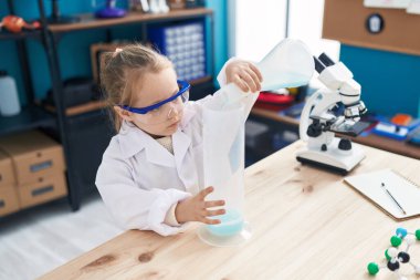 Adorable blonde girl student pouring liquid on test tube at laboratory classroom