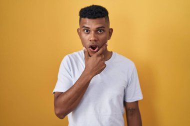 Sarı arka planda duran İspanyol genç adam, elleri çenesinde, inanamama, şaşkınlık ve şaşkınlık içinde bakıyor. 