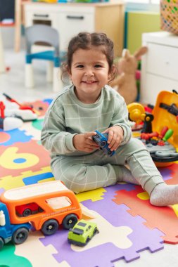 Adorable hispanic toddler playing with car toy sitting on floor at kindergarten