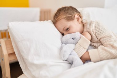 Adorable blonde girl hugging rabbit doll lying on bed at bedroom
