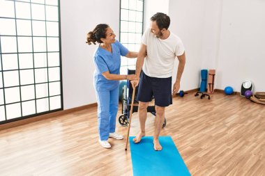 Orta yaşlı bir adam ve kadın fizyoterapi kliniğinde baston kullanarak rehabilitasyon seansı yaparken gülümsüyor.