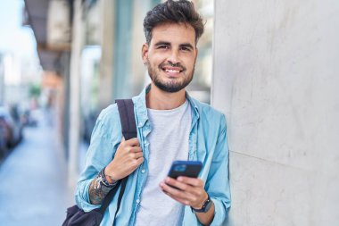 Latin asıllı genç bir öğrenci sokakta akıllı telefon kullanarak gülümsüyor.