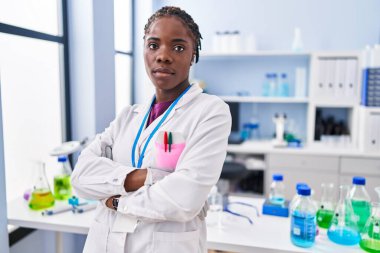 Afrika kökenli Amerikalı kadın bilim adamı üniforması giyiyor. Laboratuvarda kollarını kavuşturup el kol hareketi yapıyor.