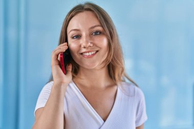 Sarışın, kendine güvenen genç bir kadın izole edilmiş mavi duvar arka planında akıllı telefondan konuşuyor.