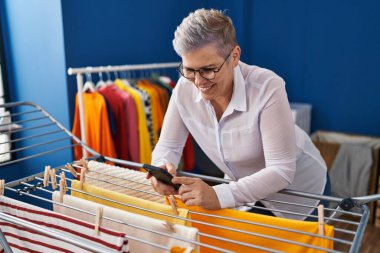 Middle age woman using smartphone hanging clothes on clothesline at laundry room