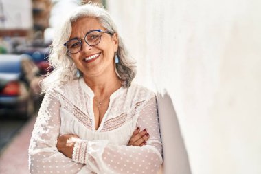Orta yaşlı kadın gülümsüyor. Kollarını kavuşturup caddede duruyor.
