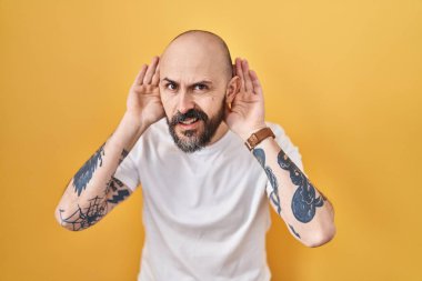 Young hispanic man with tattoos standing over yellow background trying to hear both hands on ear gesture, curious for gossip. hearing problem, deaf 