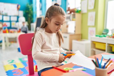 Adorable hispanic girl student sitting on table cutting paper at kindergarten