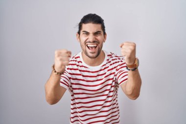 Hispanic man with long hair standing over isolated background angry and mad raising fists frustrated and furious while shouting with anger. rage and aggressive concept. 