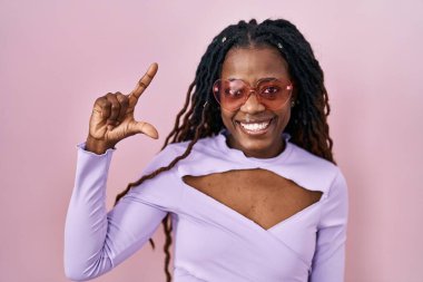 African woman with braided hair standing over pink background smiling and confident gesturing with hand doing small size sign with fingers looking and the camera. measure concept. 