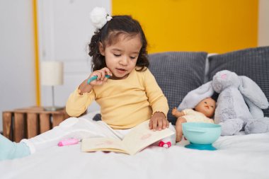 Adorable hispanic toddler sitting on bed reading book at bedroom