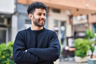 Genç Arap adam gülümsüyor. Kollarını kavuşturup caddede duruyor.
