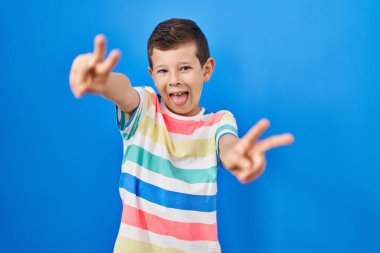 Young caucasian kid standing over blue background smiling with tongue out showing fingers of both hands doing victory sign. number two. 