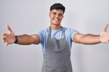 Hispanic young man wearing apron over white background looking at the camera smiling with open arms for hug. cheerful expression embracing happiness. 