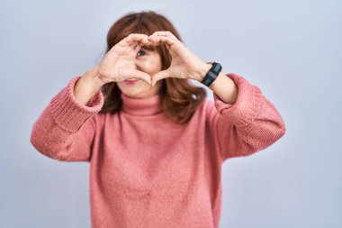 Orta yaşlı İspanyol bir kadın izole edilmiş arka planda duruyor. El ve parmaklarıyla kalp şeklinde gülümsüyor. İşaretten bakıyor. 