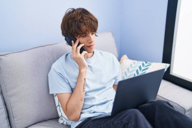 Young blond man talking on smartphone using laptop at home