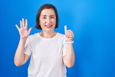 Middle age hispanic woman standing over blue background showing and pointing up with fingers number six while smiling confident and happy. 