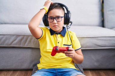 Young hispanic kid playing video game holding controller wearing headphones confuse and wondering about question. uncertain with doubt, thinking with hand on head. pensive concept. 