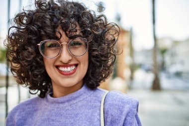 Orta Doğulu genç bir kadın gülümsüyor. Sokakta gözlük takıyor.