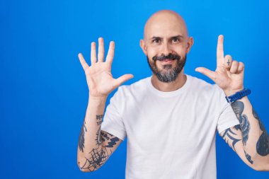 Hispanic man with tattoos standing over blue background showing and pointing up with fingers number seven while smiling confident and happy. 