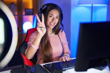Young hispanic woman playing video games smiling looking to the camera showing fingers doing victory sign. number two. 