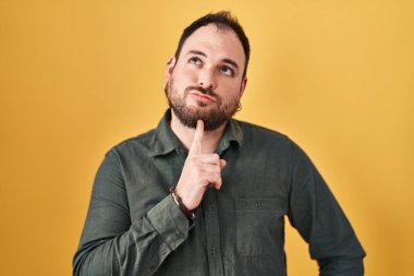 Plus size hispanic man with beard standing over yellow background thinking concentrated about doubt with finger on chin and looking up wondering 