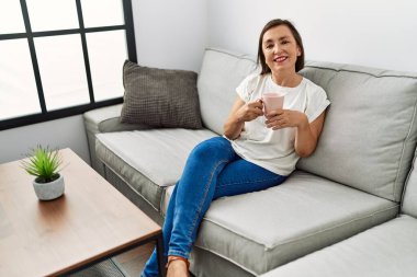 Orta yaşlı İspanyol kadın kanepede oturuyor ve evde bir fincan kahve içiyor.