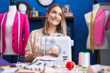 Young caucasian woman tailor smiling confident using sewing machine at sewing studio