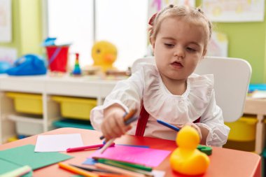 Sevimli sarışın anaokulu öğrencisi anaokulunda masaya oturmuş kağıda resim çiziyor.