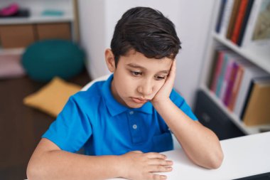 Adorable hispanic boy student using computer with tired expression at classroom