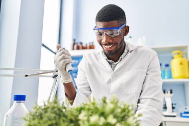 Young african american man wearing scientist uniform using pipette on plant at laboratory