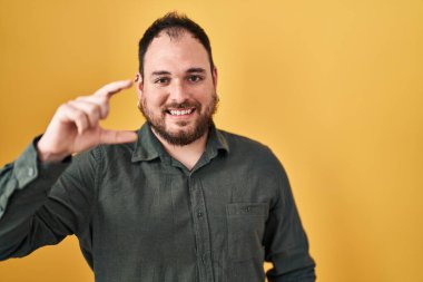 Plus size hispanic man with beard standing over yellow background smiling and confident gesturing with hand doing small size sign with fingers looking and the camera. measure concept. 