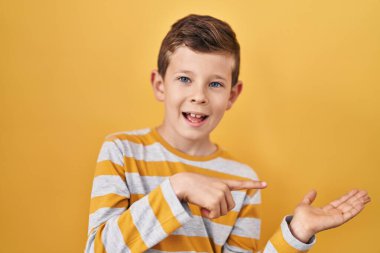 Young caucasian kid standing over yellow background amazed and smiling to the camera while presenting with hand and pointing with finger. 