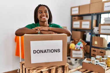 Gönüllü üniforma giyen Afro-Amerikalı genç bir kadın hayır kurumunda bağış paketine yaslanıyor.
