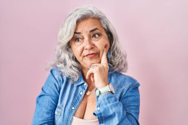 Middle age woman with grey hair standing over pink background thinking worried about a question, concerned and nervous with hand on chin 