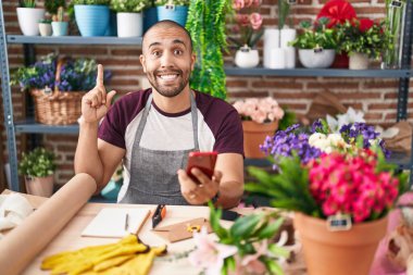Hispanic man with beard working at florist shop with smartphone smiling with an idea or question pointing finger with happy face, number one 