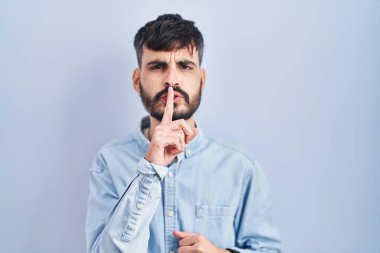 Young hispanic man with beard standing over blue background asking to be quiet with finger on lips. silence and secret concept. 