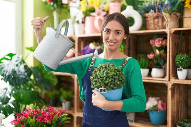 Young beautiful hispanic woman florist smiling confident watering plant at flower shop