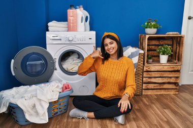 Young hispanic woman doing laundry smiling doing phone gesture with hand and fingers like talking on the telephone. communicating concepts. 